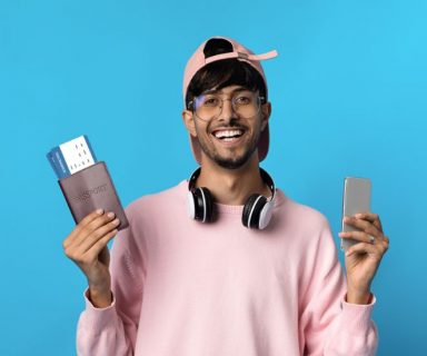 A young man holding a passport, plane tickets and a cell phone. He is standing against a light blue background.