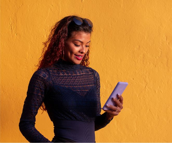 A woman in a long sleeved blue top holding a cell phone, standing against a dark yellow wall.