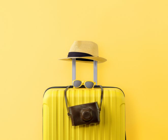 A yellow suitcase against a yellow wall. There is a beach hat and camera bag on top of it.